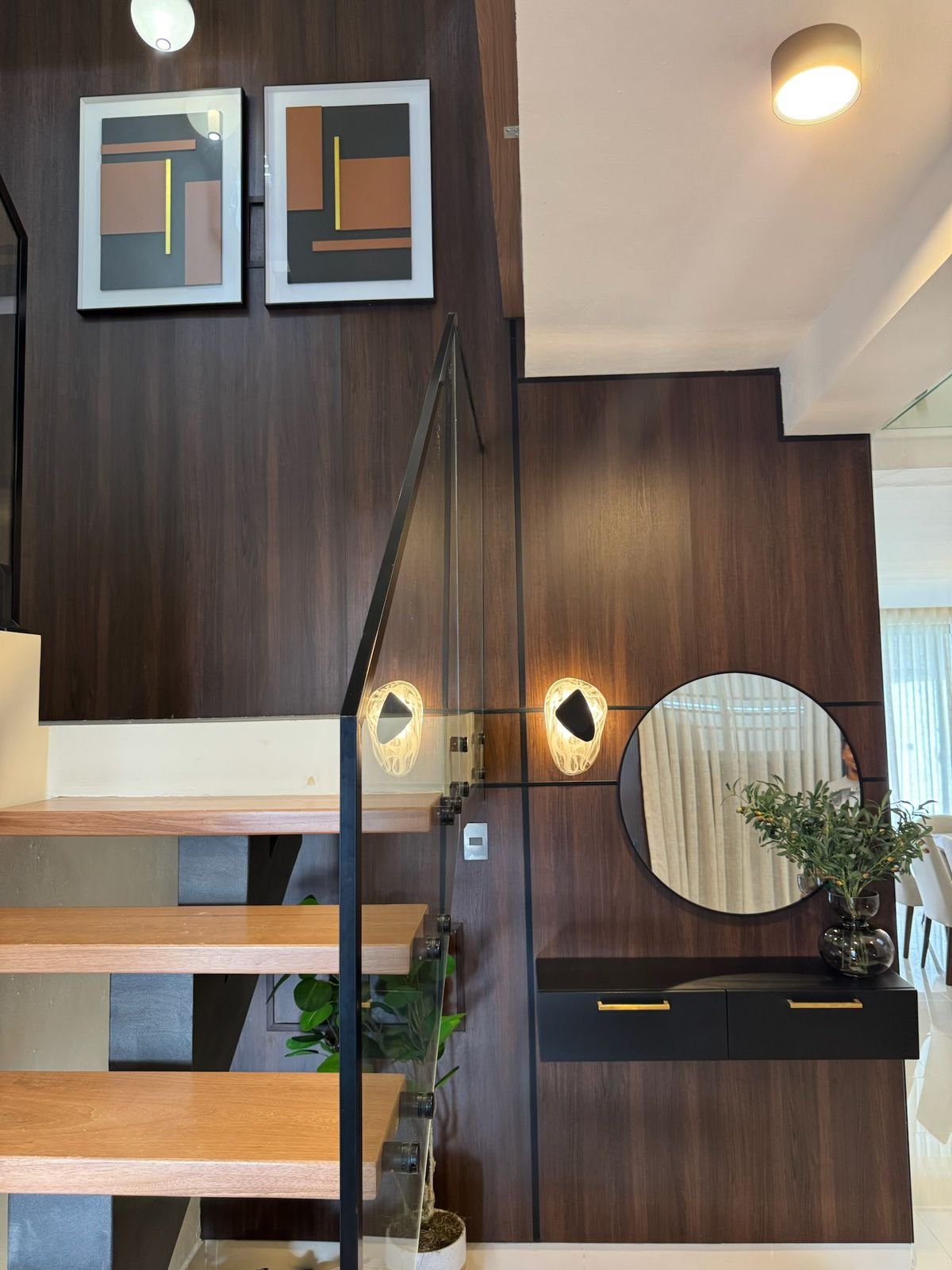 Modern interior with dark wood wall featuring framed artwork, round mirror, pendant lights, and wooden staircase with plants