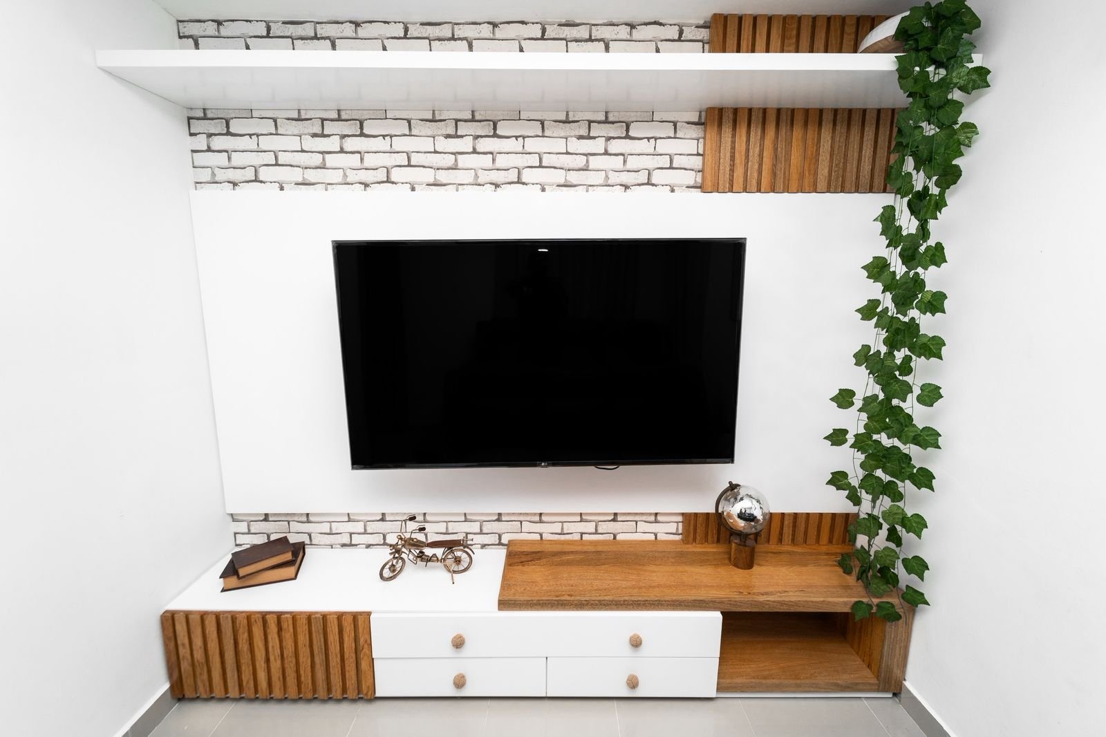 Modern entertainment center with wall-mounted TV, wooden shelving, white storage cabinets, and climbing ivy plant against white brick wall