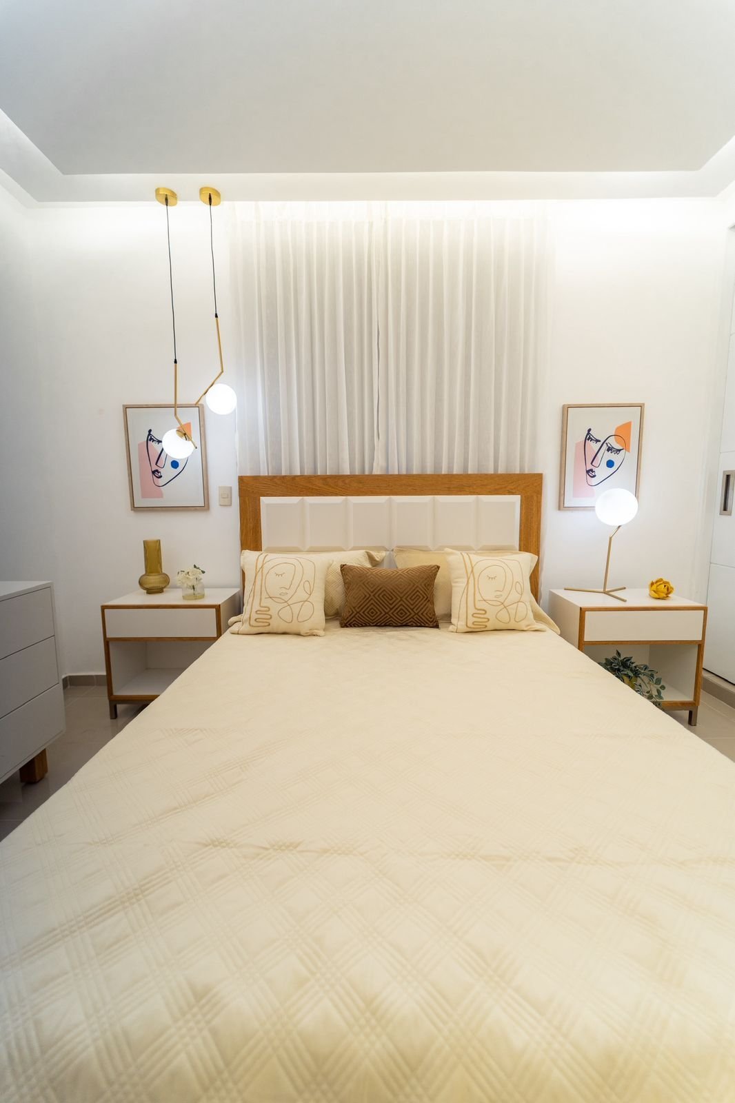 Modern bedroom with wooden bed frame, cream bedding, wooden nightstands, hanging pendant lights, and minimalist white walls with curtains