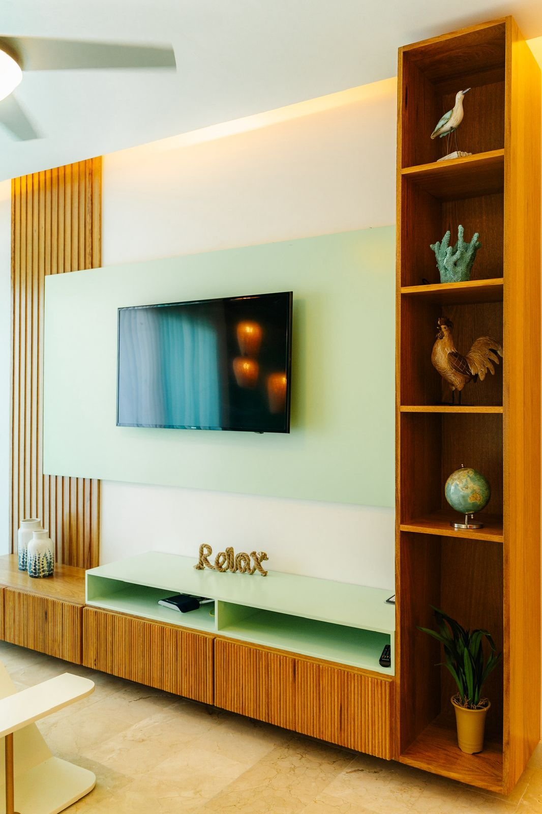 Modern living room with wall-mounted TV, wooden entertainment unit with Relax sign, and tall wooden shelving displaying decorative items