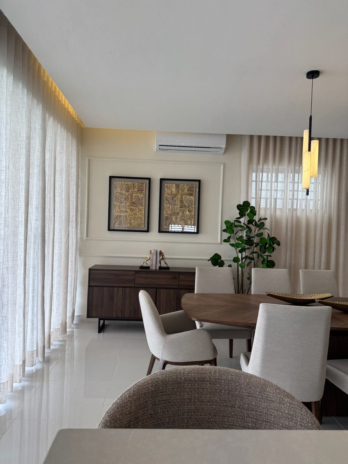Modern dining room with wood table, white upholstered chairs, dark credenza, framed wall art, floor-to-ceiling curtains, and potted plant
