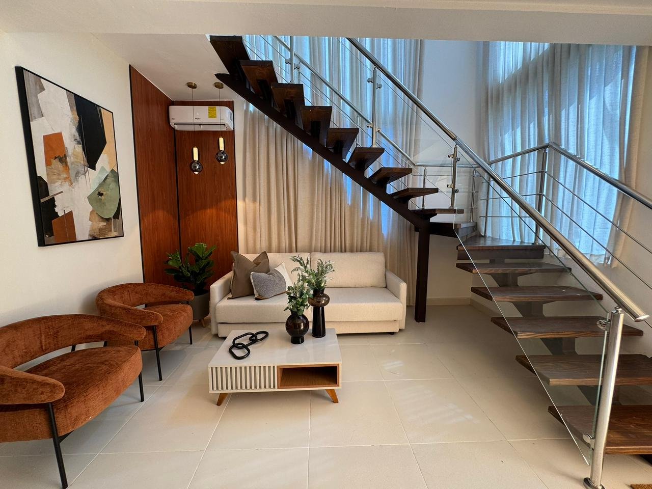 Modern living room with cream-colored couch, brown chairs, wooden staircase, potted plants, and floor-to-ceiling windows with sheer curtains