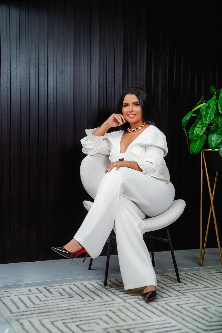 Woman in white blazer and pants sitting on chair against dark striped wall with potted plant nearby