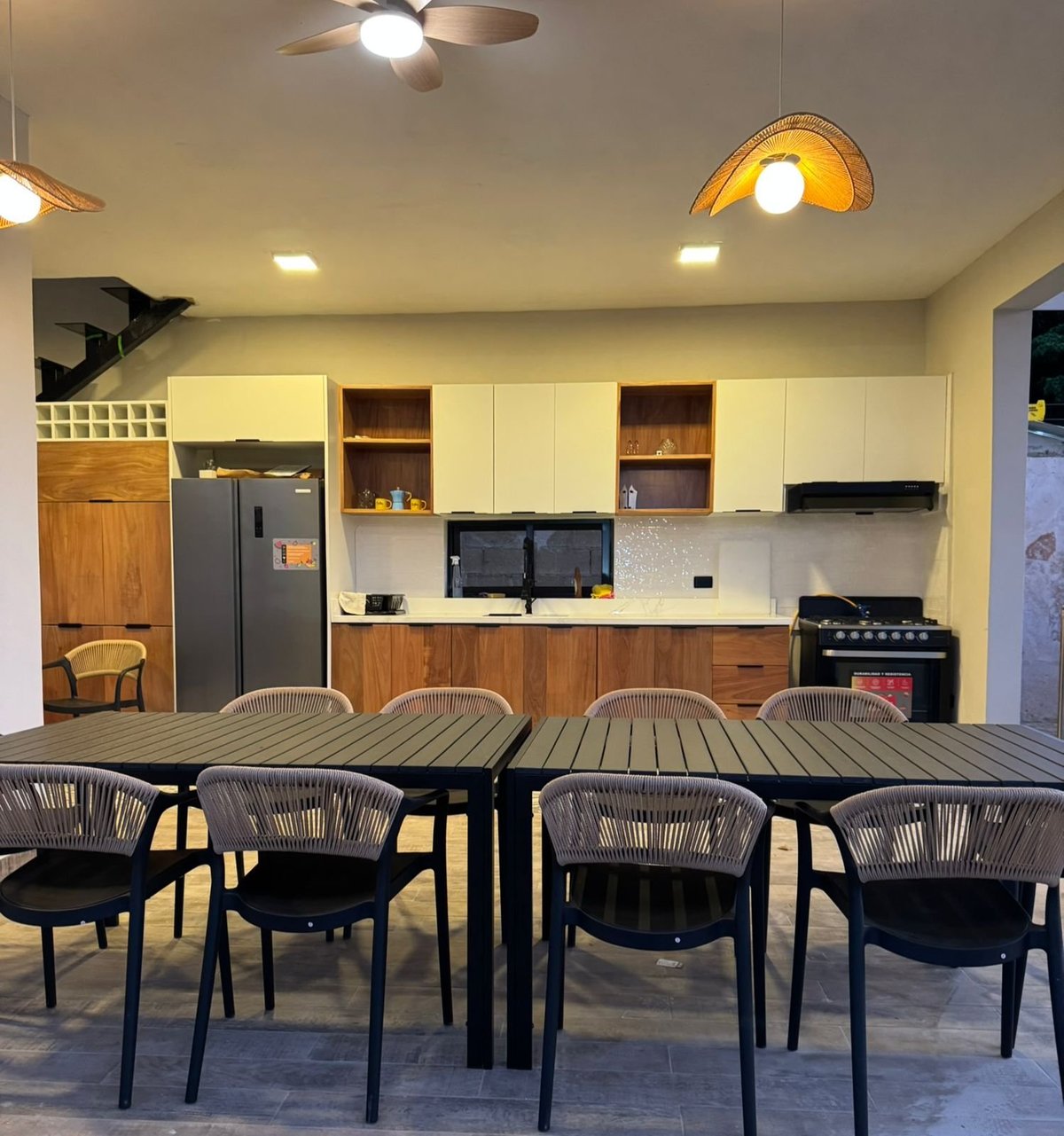 Modern kitchen and dining area with black chairs around a wooden table, cream and wood cabinetry, stainless steel appliances, and ceiling fans with warm lighting