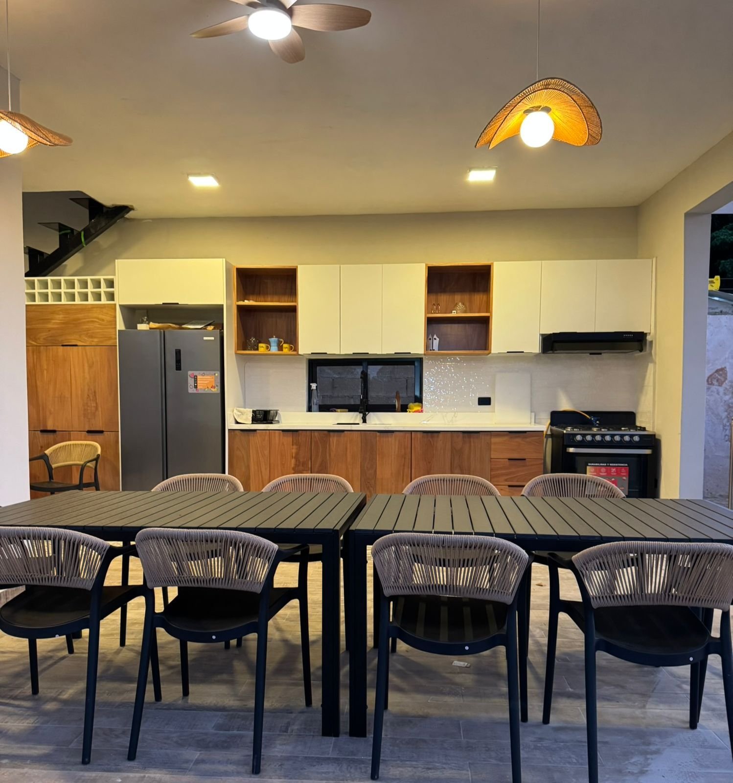 Modern kitchen and dining area with black chairs around a wooden table, cream and wood cabinetry, stainless steel appliances, and ceiling fans with warm lighting