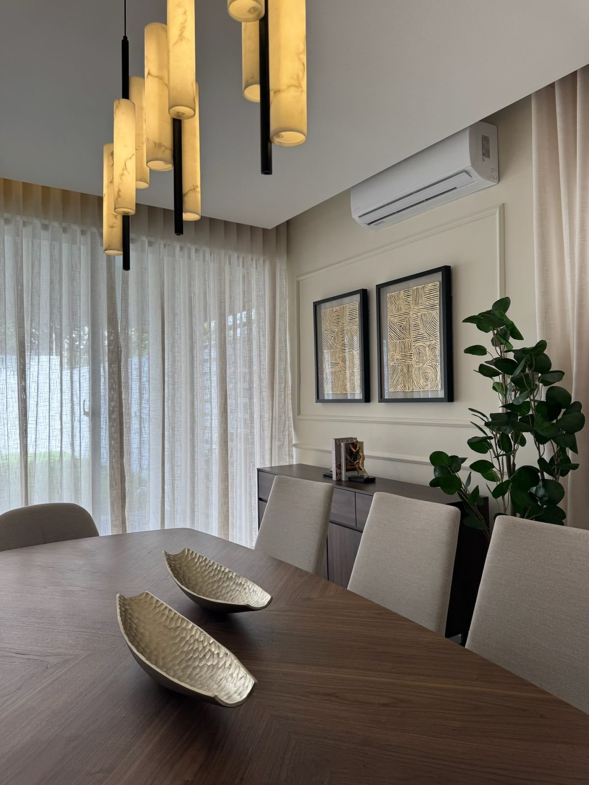 Modern dining room with wooden table, upholstered chairs, gold pendant lighting, framed wall art, potted plant, and sheer curtains